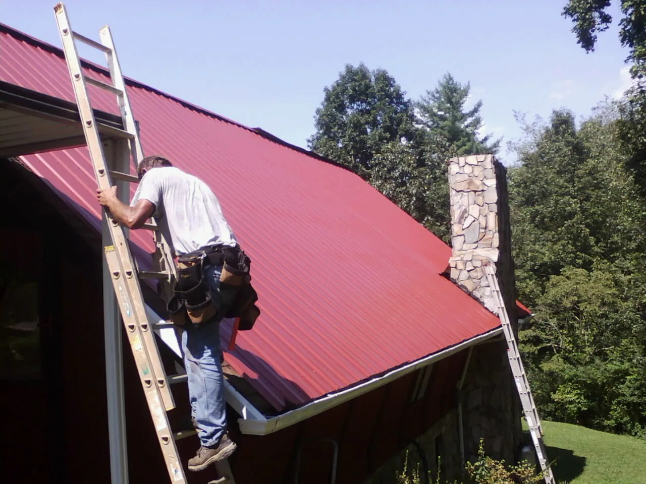 Roofer on a ladder beside a red metal roof and stone chimney — Soffit & Fascia Repair by Roofing Brothers in Candler